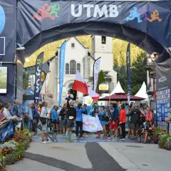 Uczestnicy Ultra-Trail du Mont-Blanc świętują pod łukiem startowym. W tle kościół i tłum widzów.