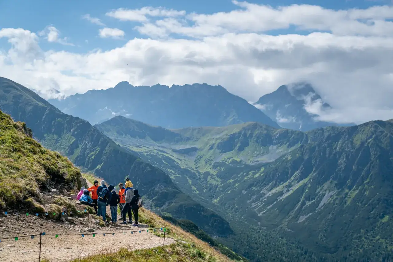 Grupa turystów odpoczywa na szlaku z widokiem na Tatry. Kasprowy Wierch gdzie jest, widać skaliste zbocza i zielone doliny.